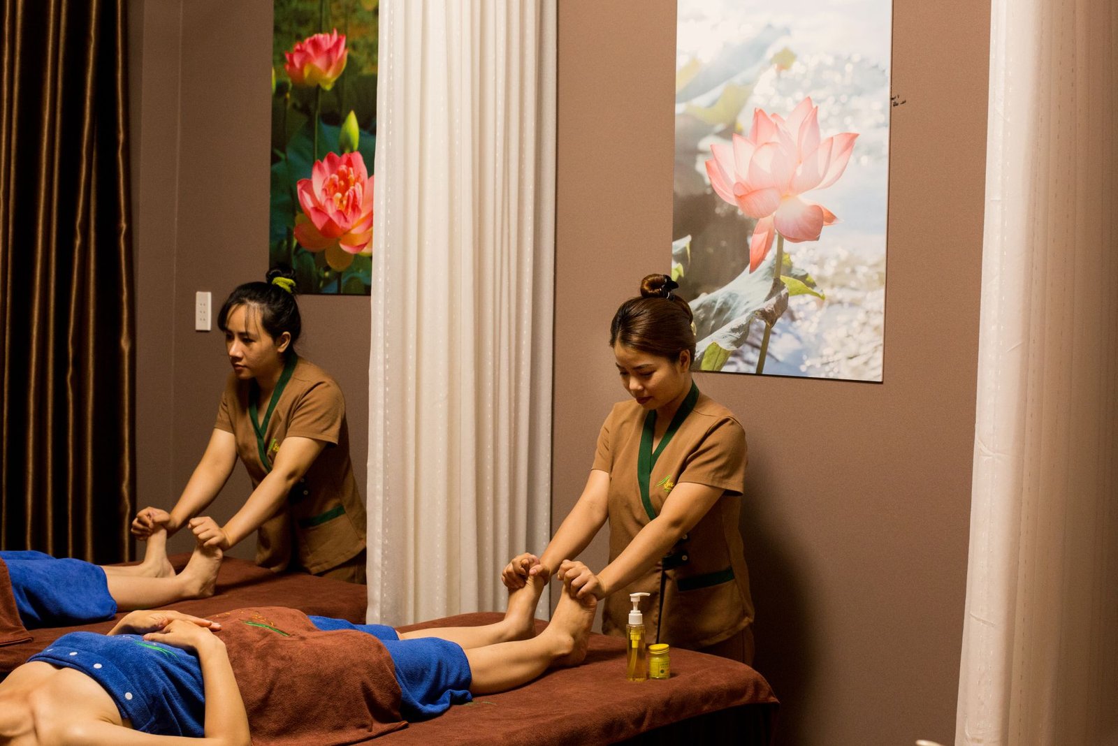 Hai nhân viên spa đang thực hiện mát-xa chân cho khách hàng trong phòng có tranh hoa sen.