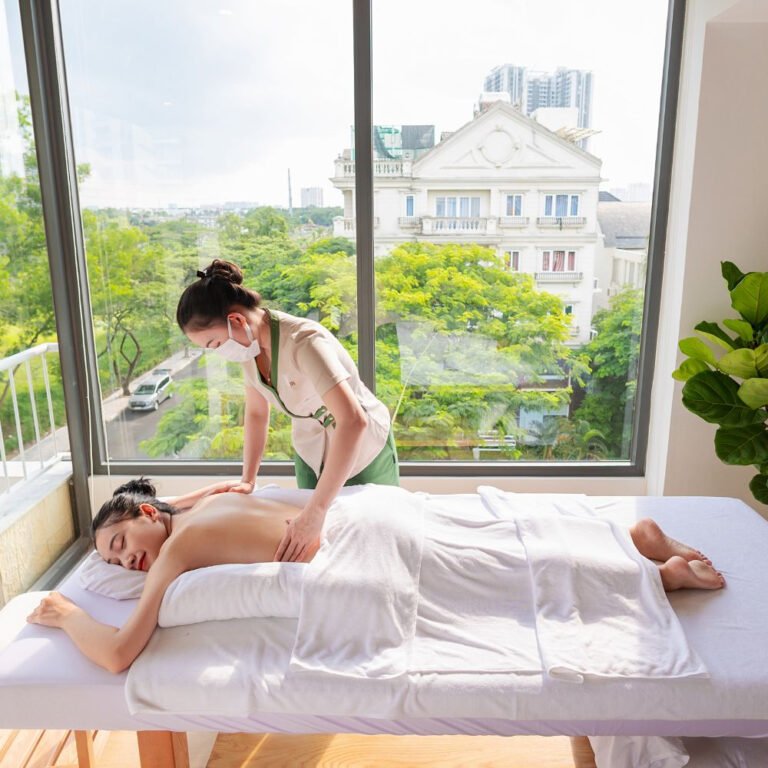 Nhân viên spa thực hiện massage thư giãn cho khách hàng bên cửa sổ lớn nhìn ra quang cảnh xanh tươi.