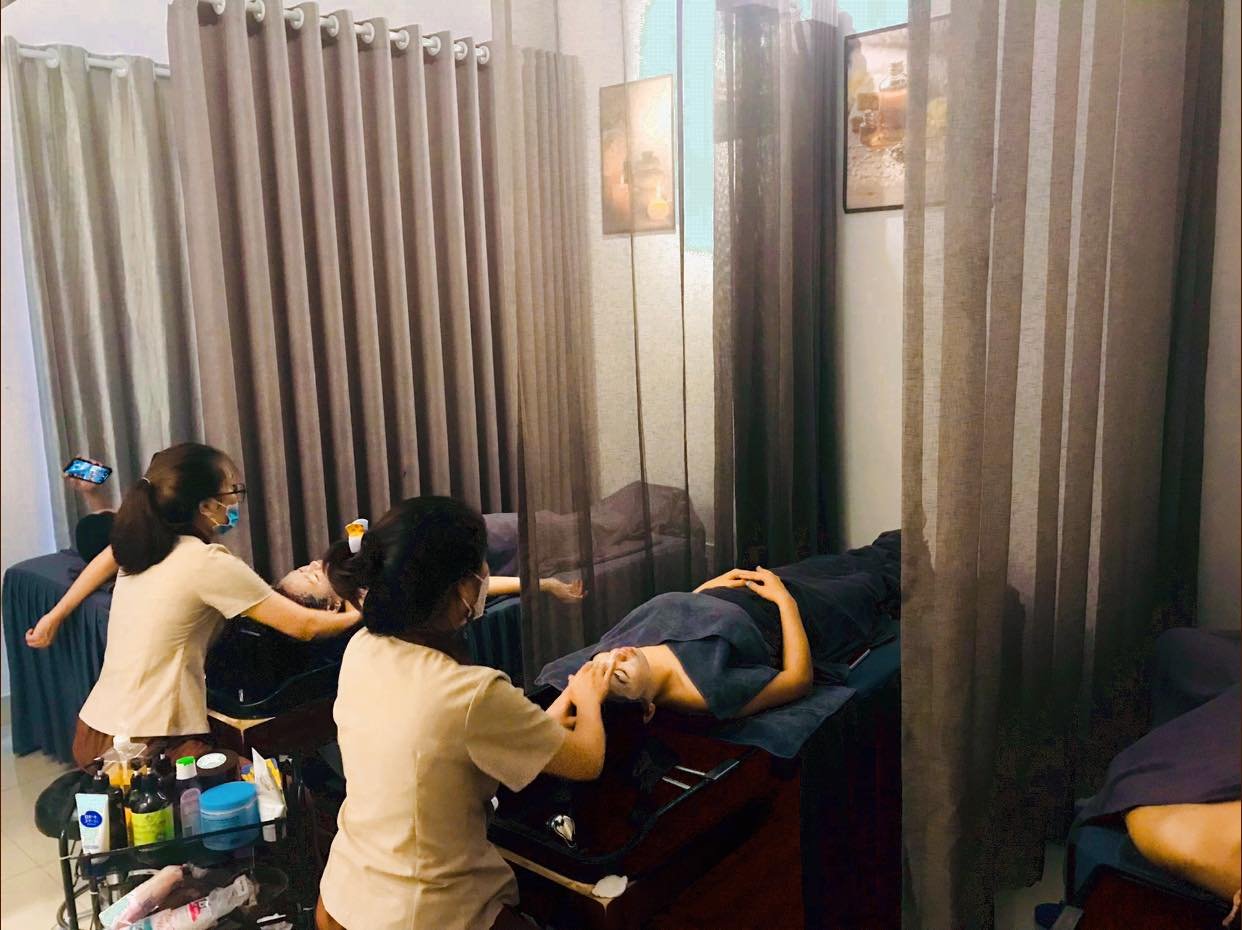 Hai người đang chăm sóc da mặt trong một căn phòng spa với rèm che và nhiều sản phẩm làm đẹp.