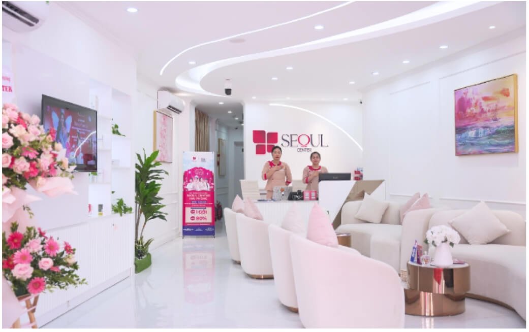 Nội thất phòng chờ sang trọng của trung tâm thẩm mỹ với hoa và tranh trang trí, hai nhân viên lễ tân làm việc.