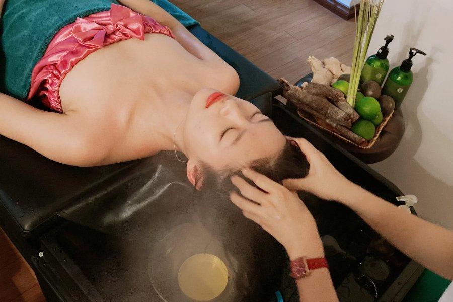 Người phụ nữ được xông hơi tóc tại spa, với các chai dầu gội thảo dược bên cạnh.