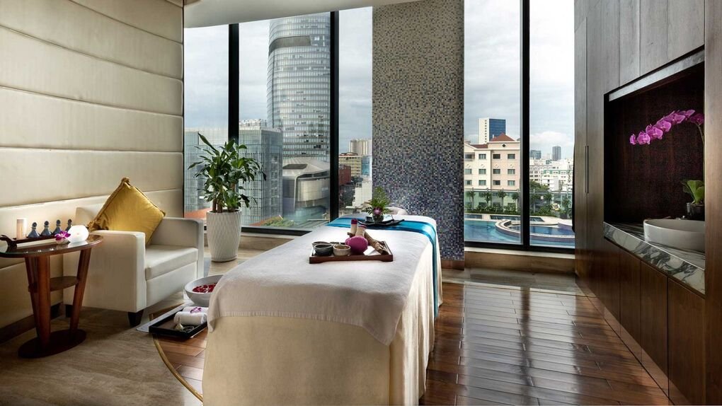 Phòng spa sang trọng với bàn massage, cây xanh, và tầm nhìn thành phố qua cửa sổ kính lớn.