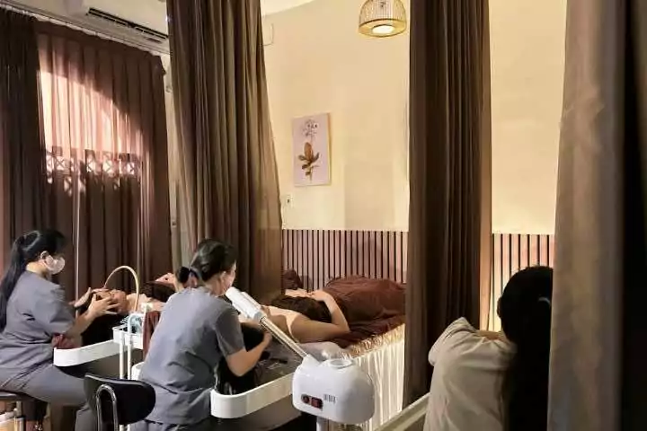Nhân viên spa chăm sóc da mặt cho khách hàng trong không gian thư giãn với rèm màu nâu.
