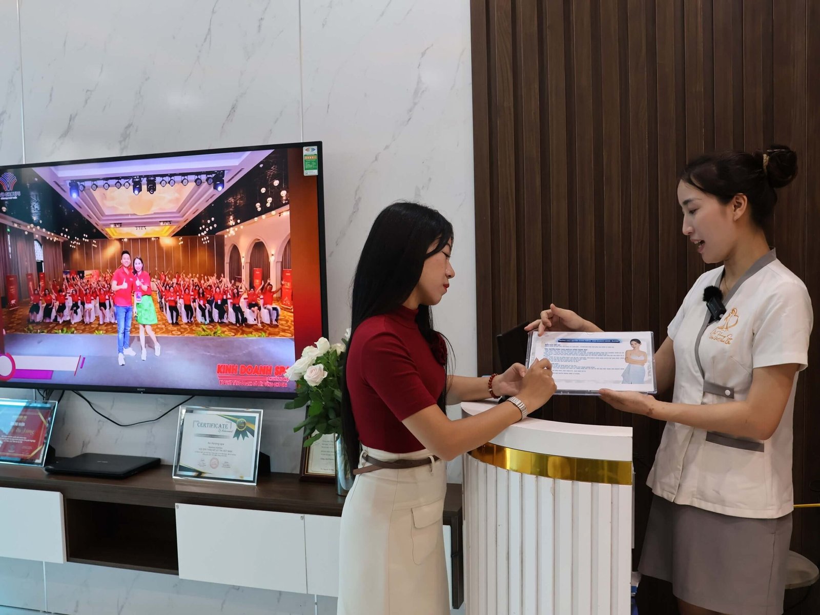 Hai người đang trò chuyện tại quầy lễ tân, một người cầm tài liệu. Trong phòng có màn hình TV và chứng nhận.