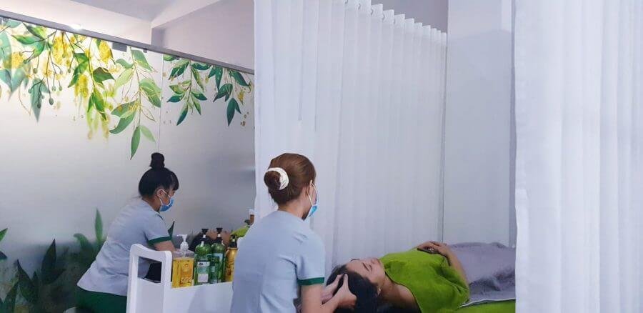 Hai nhân viên chăm sóc khách hàng trong spa, một người nằm thư giãn trên giường với khăn phủ, không gian trang trí cây lá.