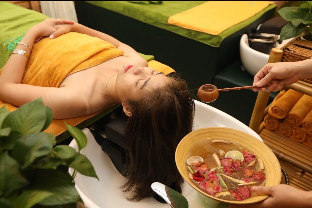 Người phụ nữ nằm thư giãn trong spa, chuẩn bị được gội đầu bằng nước thảo dược trong bát.