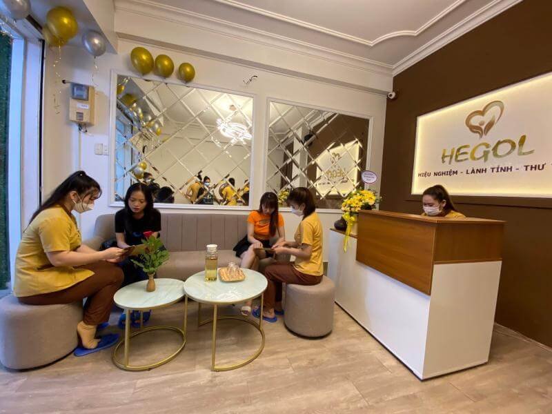 Nhân viên và khách hàng tại spa Hegol, không gian với ghế sofa, bàn tròn và trang trí bóng bay vàng.