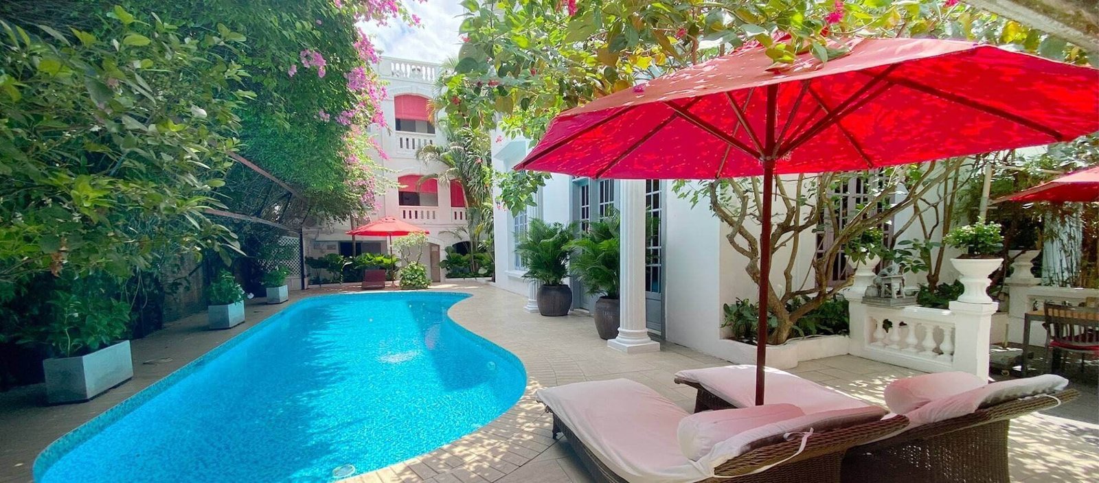 Hồ bơi ngoài trời xanh mát với ghế dài và ô đỏ, bao quanh bởi cây cối xanh tươi trong khuôn viên biệt thự.