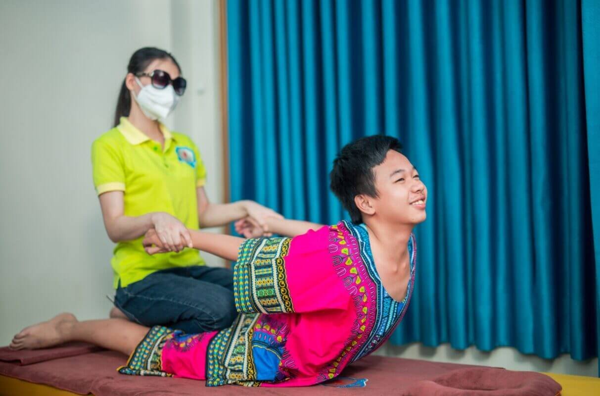 Người được mát-xa Thái truyền thống, mặc áo sắc màu, thư giãn trong phòng có rèm xanh.