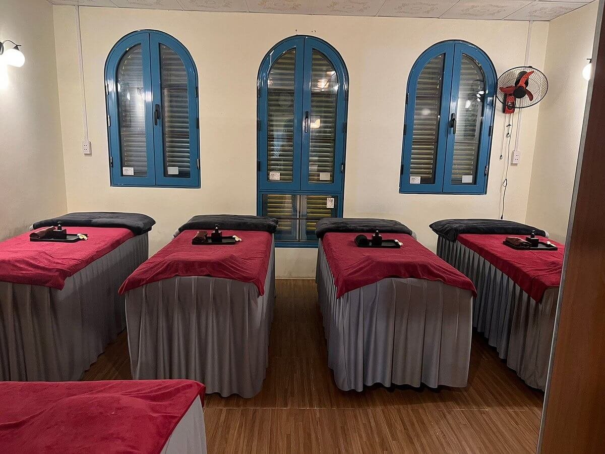 Phòng massage với giường phủ khăn đỏ, cửa sổ xanh, ánh sáng ấm áp, tạo không gian thư giãn và thoải mái.