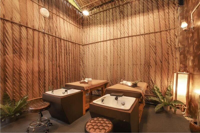 Phòng spa thư giãn với giường massage đôi và bồn rửa, trang trí bằng cây xanh và đèn ấm áp.