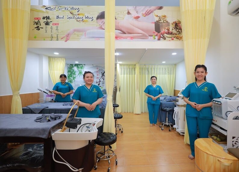Bốn nhân viên spa đứng trong phòng trị liệu, mặc đồng phục xanh, chuẩn bị đón khách.