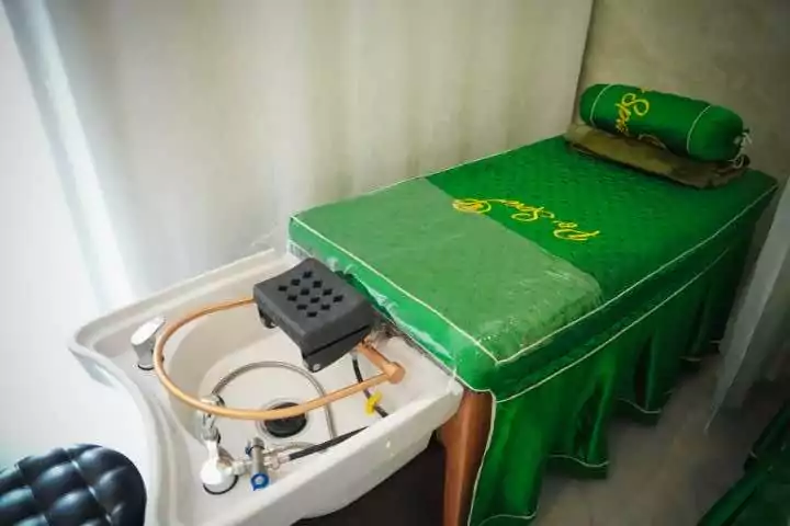 Giường massage với tấm phủ màu xanh lá cây và bồn rửa bên cạnh trong phòng spa thư giãn.