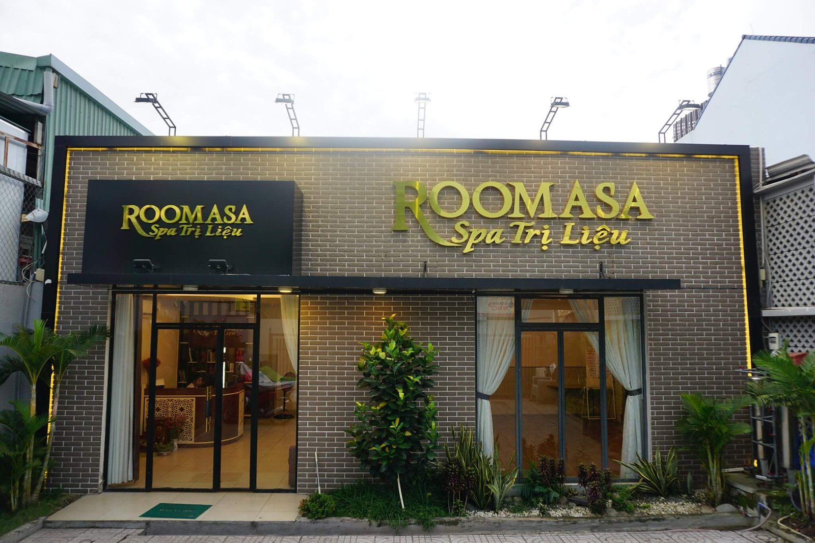 Mặt tiền spa Roomasa với bảng hiệu Spa Trị Liệu và cửa kính, trang trí cây xanh.