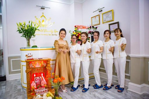 Nhân viên spa mặc đồng phục trắng tạo dáng trước quầy lễ tân với trang trí hoa và giấy chứng nhận trên tường.