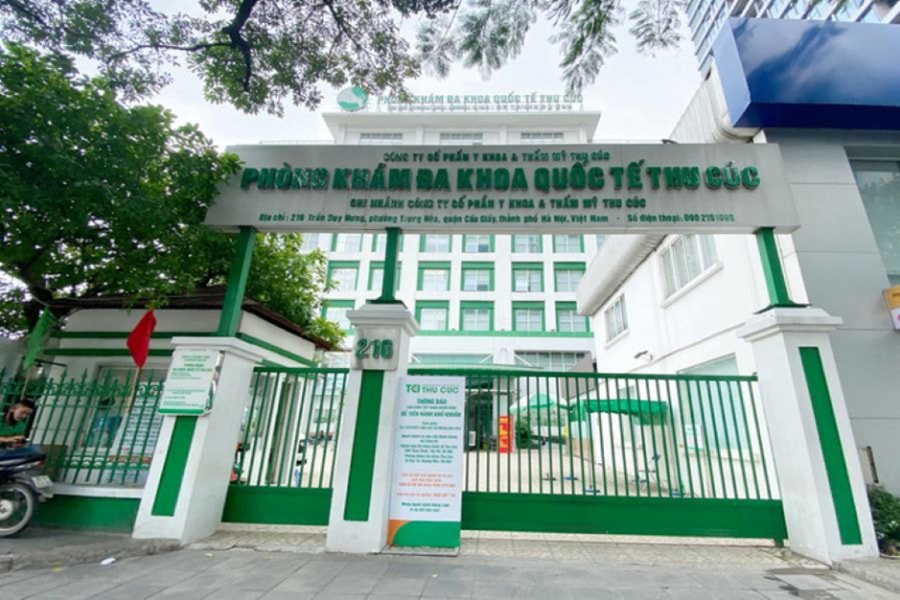 Cổng vào của một phòng khám đa khoa màu trắng và xanh lá cây, với biển hiệu và cây xanh xung quanh.