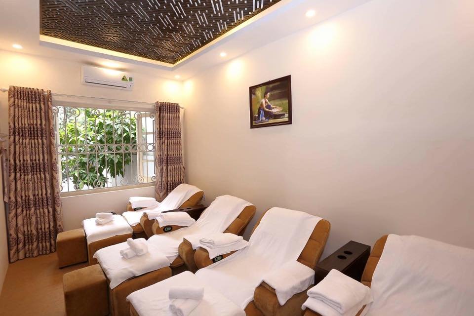 Phòng spa với ghế massage, khăn trắng, ánh sáng dịu nhẹ và tranh treo tường, tạo không gian thư giãn.