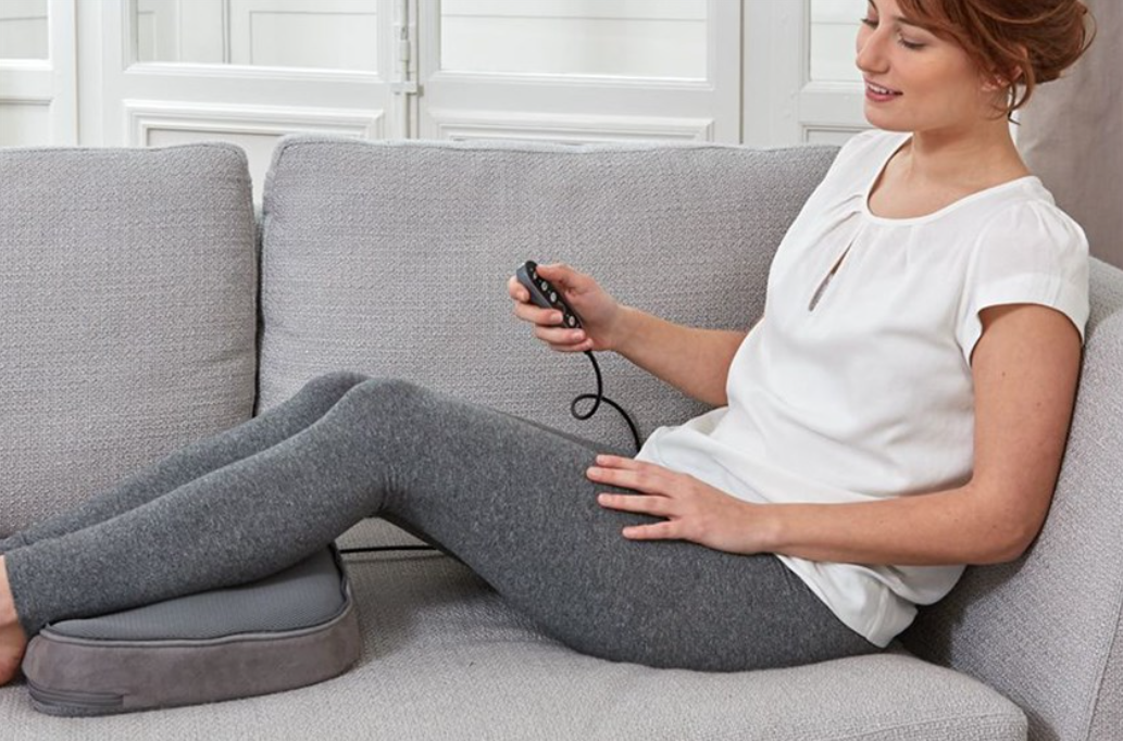 Người ngồi trên ghế sofa, cầm điều khiển và đặt chân trên gối massage màu xám.