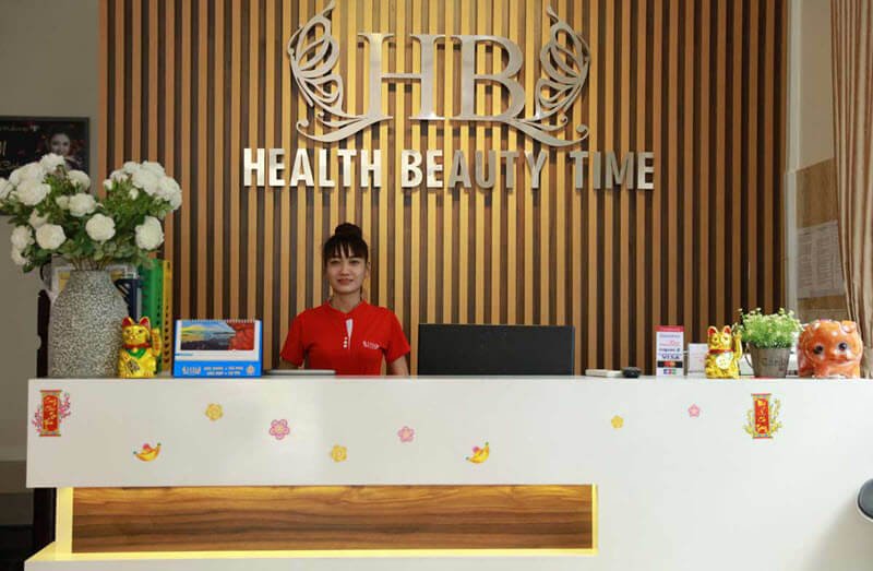 Quầy lễ tân với nhân viên đứng trước biển hiệu "Health Beauty Time," trang trí hoa và vật phẩm may mắn.