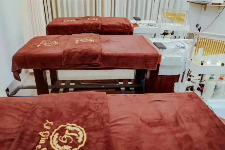 Ba giường mát-xa có khăn trải màu nâu, trong một phòng spa với rèm trắng và các thiết bị chăm sóc da.