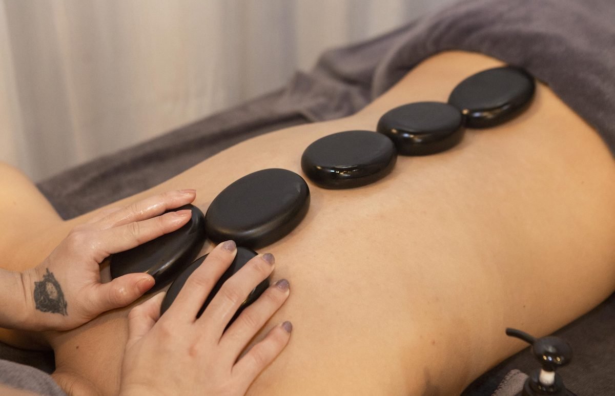 Người đang thực hiện massage bằng đá nóng trên lưng, có hình xăm trên tay và khăn phủ dưới cơ thể.