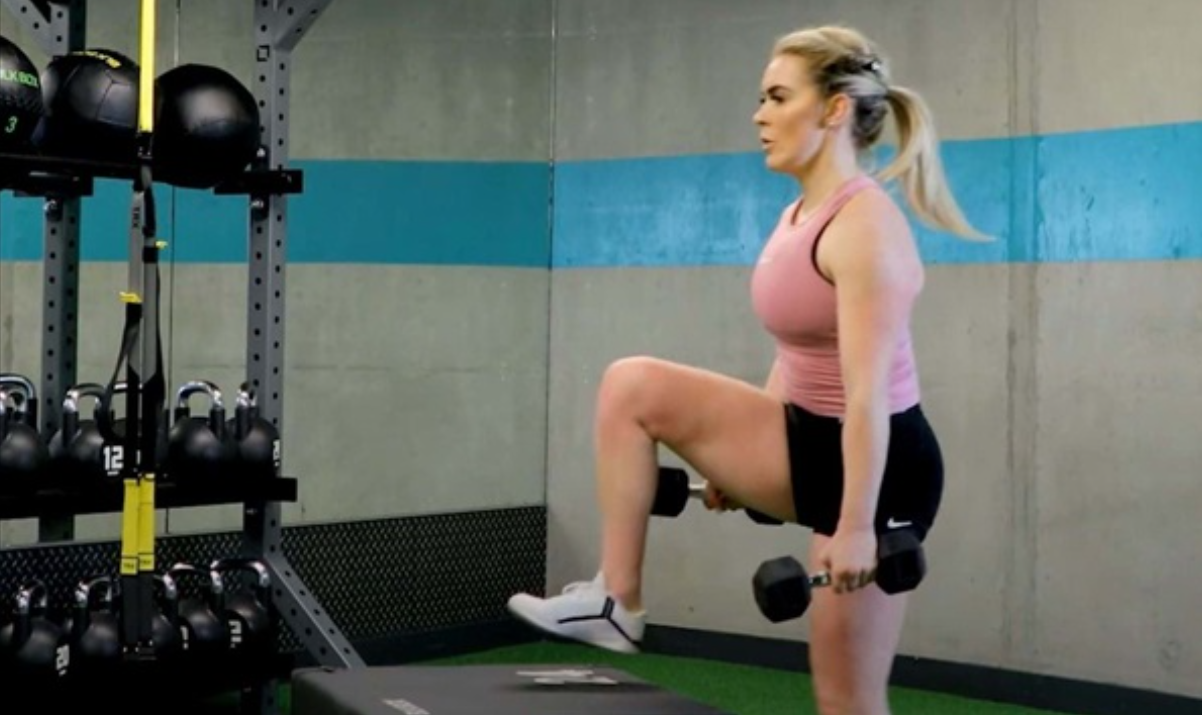 Người phụ nữ đang thực hiện bài tập với tạ tay trong phòng gym, xung quanh có nhiều thiết bị tập luyện.