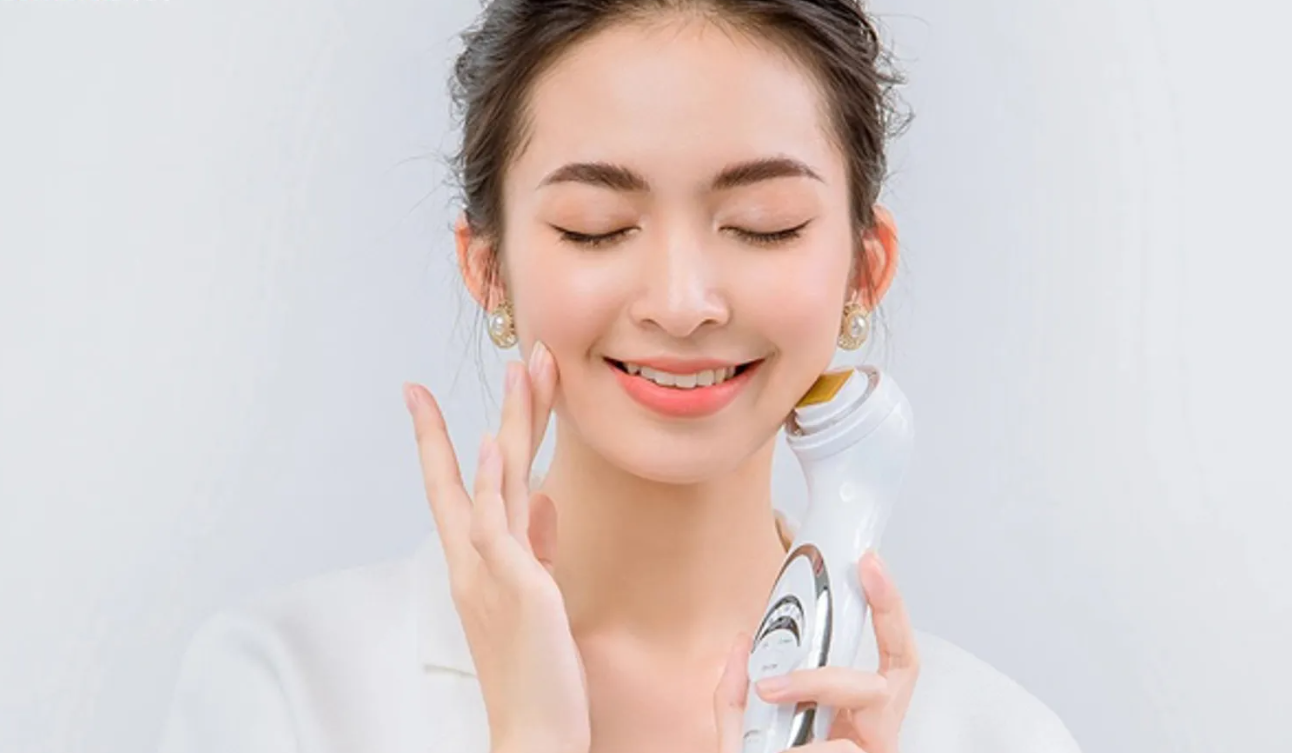 Người phụ nữ mỉm cười, nhắm mắt, tay cầm máy massage mặt, mặc áo trắng, nền trắng.