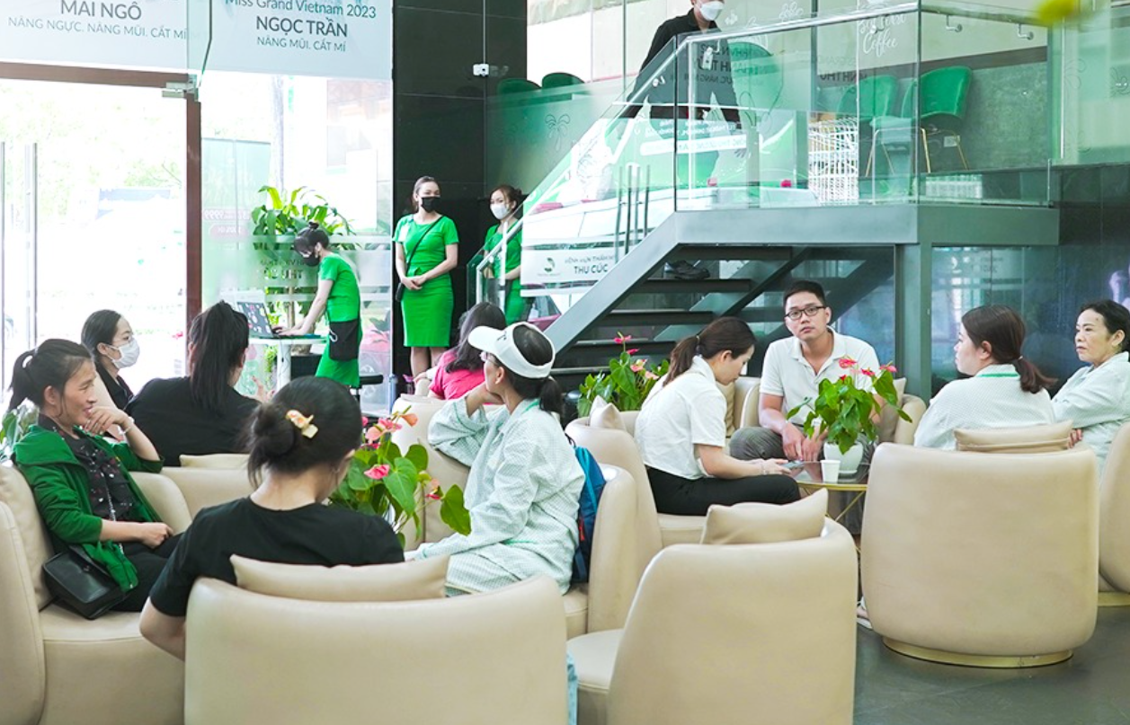 Mọi người trò chuyện trong phòng chờ với ghế sofa và cây cảnh, nhân viên mặc đồng phục xanh đứng gần cầu thang.