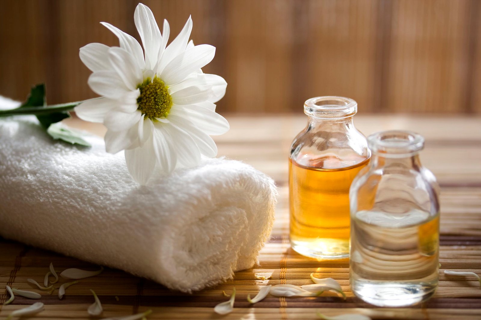 Khăn tắm trắng cuộn tròn, hoa trắng và hai lọ dầu massage trên nền gỗ, tạo cảm giác thư giãn.
