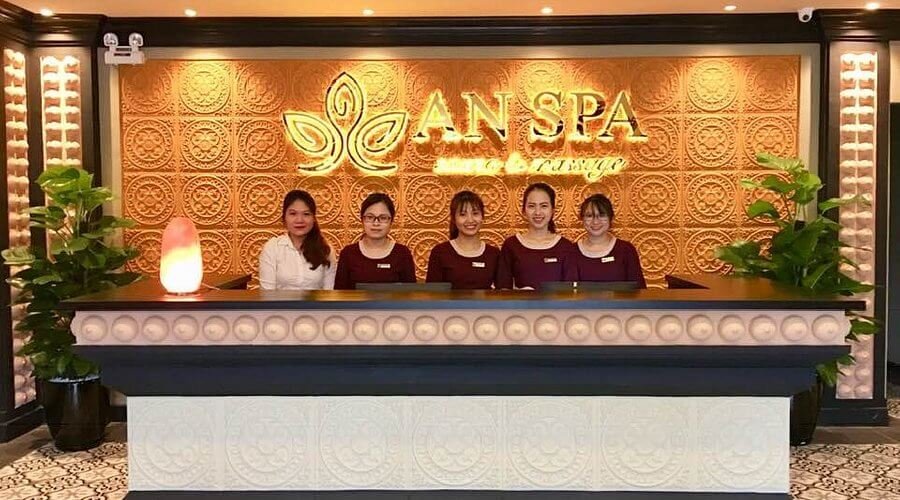 Nhóm người đứng sau quầy lễ tân trong spa, nền trang trí với logo và họa tiết hoa văn.