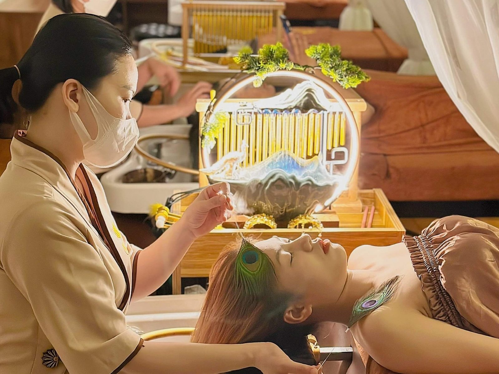 Người phụ nữ đeo khẩu trang đang chăm sóc sắc đẹp cho khách hàng nằm thư giãn trên giường spa.