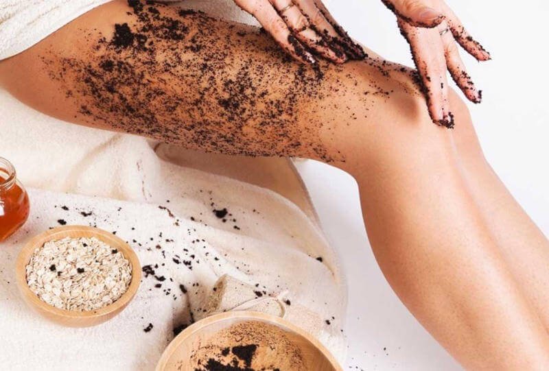 Người đang tẩy tế bào chết trên chân bằng bã cà phê, bên cạnh có bát yến mạch và mật ong.
