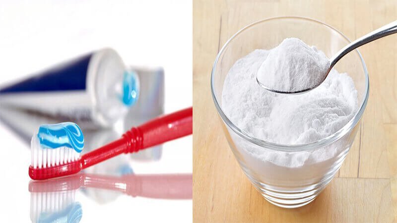 Bàn chải đánh răng với kem đánh răng bên trái và bát đựng bột trắng với muỗng bên phải trên mặt bàn.