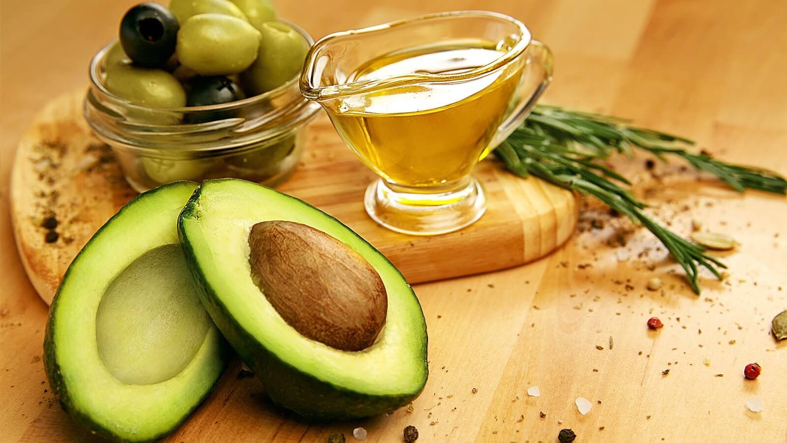 Quả bơ cắt đôi, chén dầu và ô liu trên bàn gỗ, xung quanh là các loại gia vị và thảo mộc.