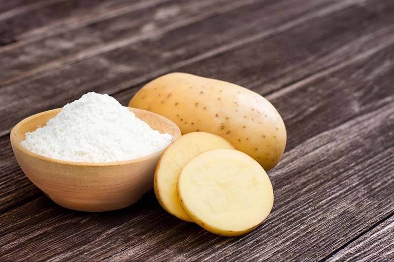 Củ khoai tây nguyên vẹn và cắt lát, bên cạnh là bát bột trắng, trên nền gỗ nâu.