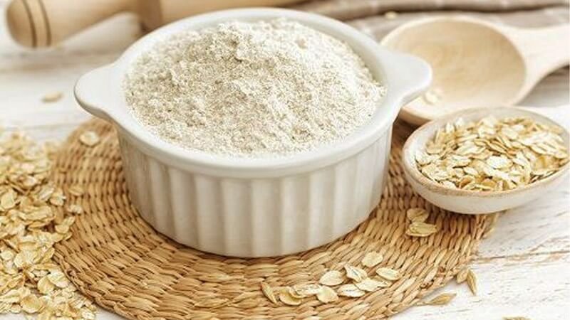 Bột yến mạch trong bát trắng, đặt trên tấm lót mây, cạnh đó là thìa gỗ và yến mạch nguyên hạt.