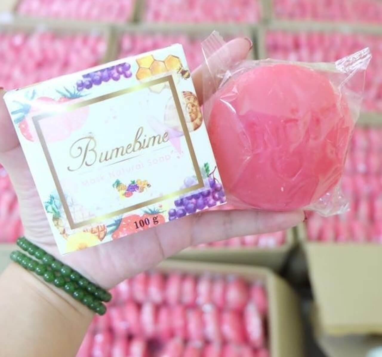 Bàn tay cầm một hộp xà phòng và một bánh xà phòng màu hồng, phía sau là nhiều hộp xà phòng tương tự.
