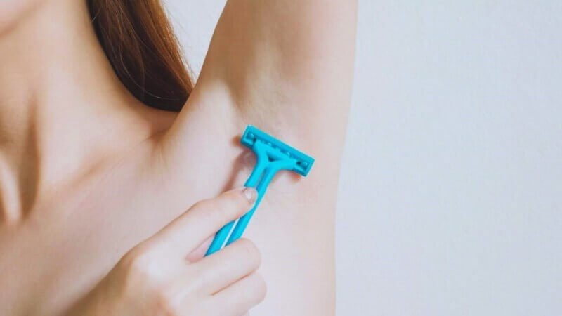Người đang dùng dao cạo màu xanh để cạo lông dưới cánh tay trên nền tường trắng.