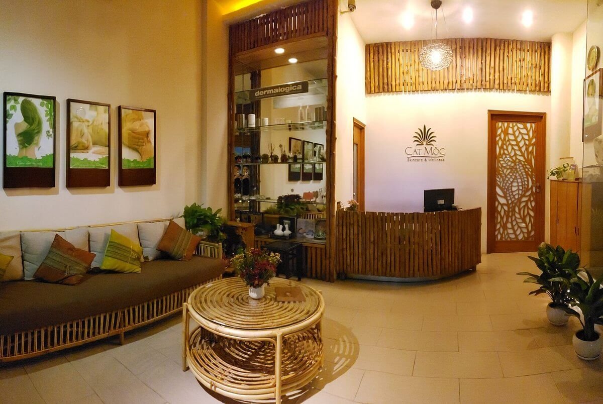 Phòng chờ spa với bàn ghế bằng tre, tường trang trí hình ảnh và quầy lễ tân bằng gỗ, ánh sáng ấm áp.