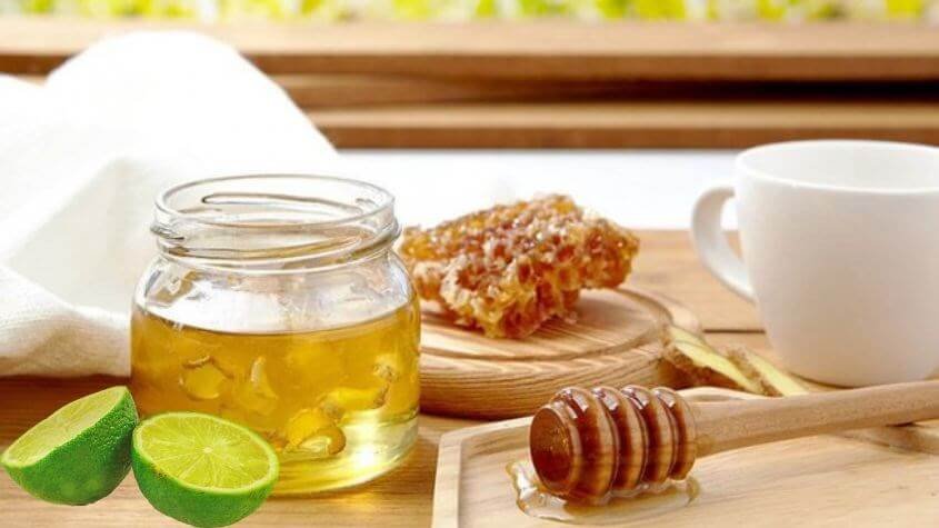 Hũ mật ong, lát chanh, và dụng cụ múc mật ong trên bàn gỗ, bên cạnh là một tách trà trắng.