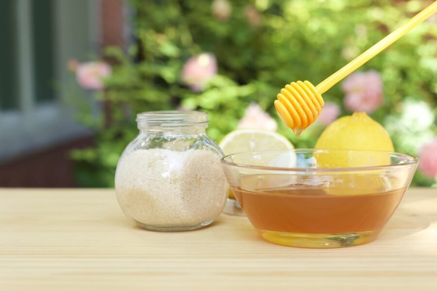 Bát mật ong với thìa khuấy, cạnh đó là lọ đường và quả chanh trên bàn gỗ, nền cây xanh mờ.