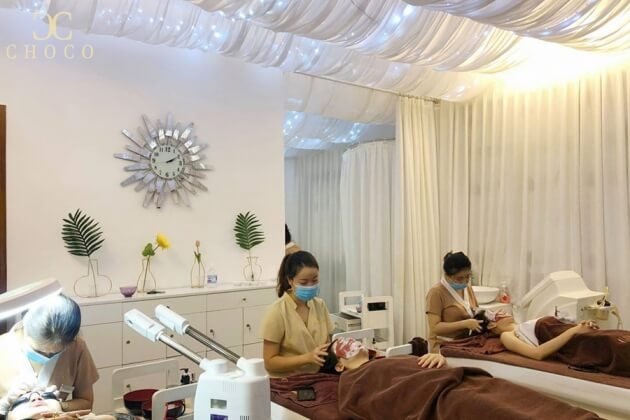 Một phòng spa với hai nhân viên đang chăm sóc da mặt cho khách hàng, trang trí tường với đồng hồ và cây cảnh.
