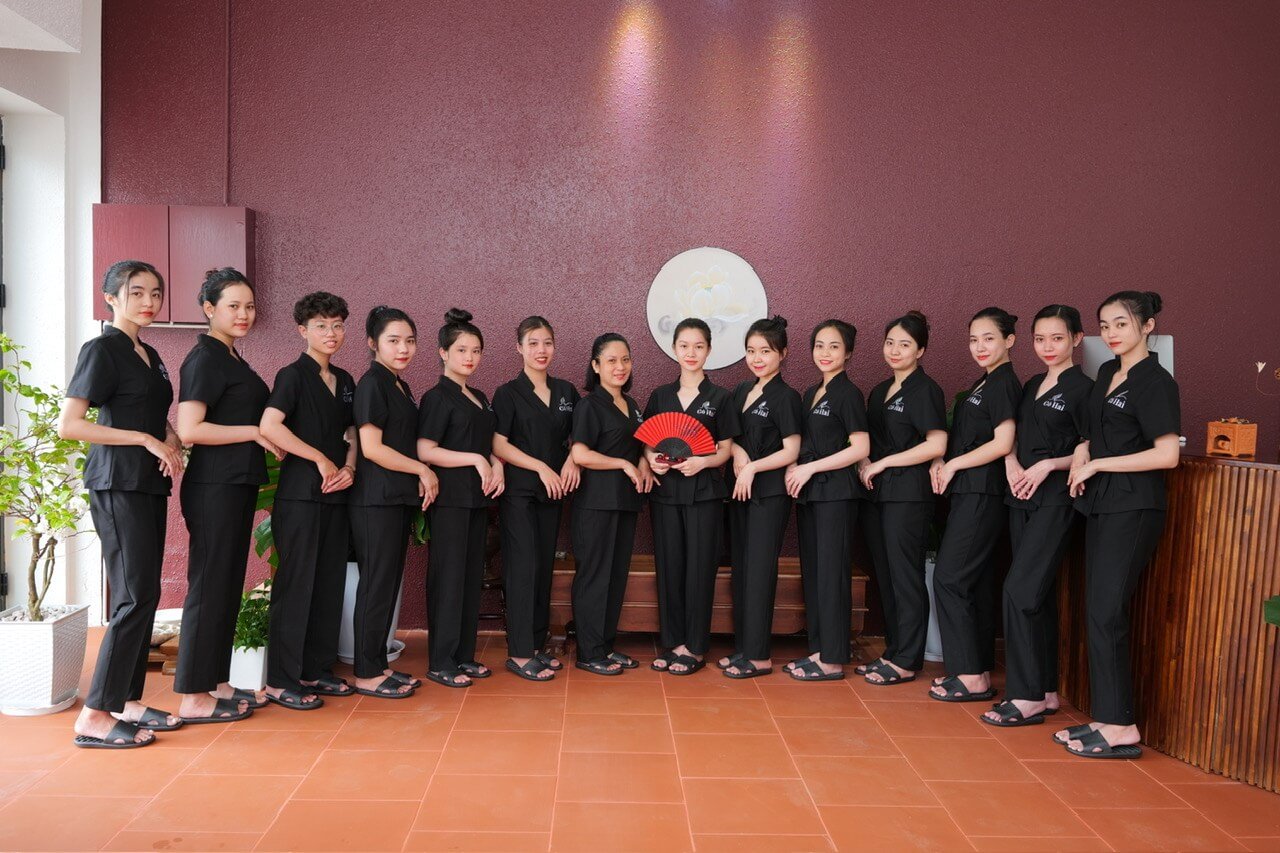 Nhóm người đứng thành hàng, mặc đồng phục đen, trên nền tường màu đỏ, một người cầm quạt đỏ.