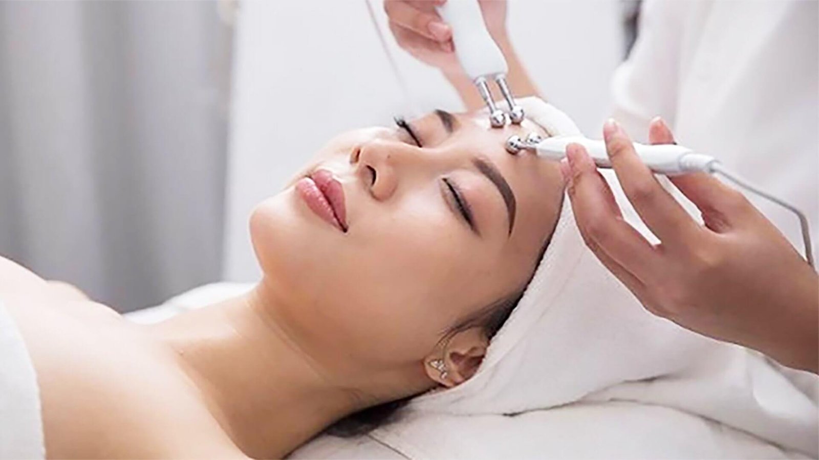 Người nằm thư giãn, được chăm sóc da mặt bằng dụng cụ chuyên dụng trong spa, khăn quấn quanh đầu.