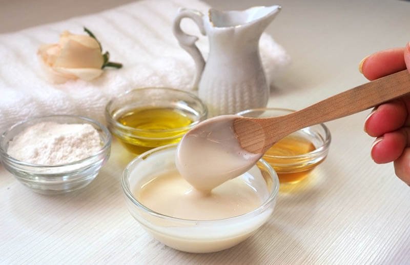 Bàn tay cầm thìa gỗ đang khuấy hỗn hợp trong bát, bên cạnh có các bát nhỏ đựng dầu, bột và chất lỏng khác.
