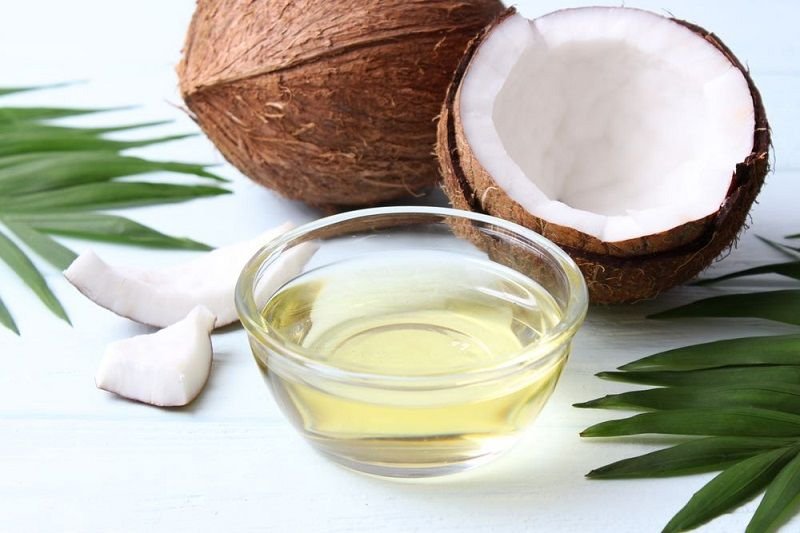 Nửa quả dừa, dầu dừa trong bát thủy tinh nhỏ, lát dừa và lá xanh trên nền trắng.