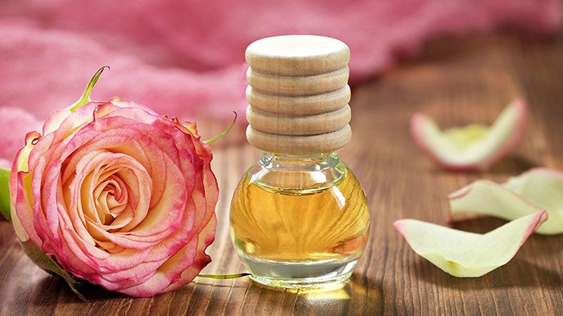 Chai tinh dầu và hoa hồng trên bàn gỗ, xung quanh có cánh hoa rơi.