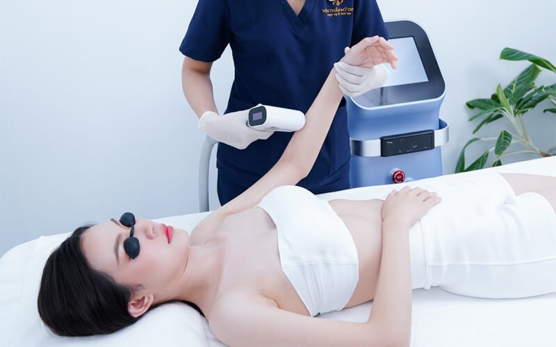 Người phụ nữ nằm trên giường trong phòng thẩm mỹ, được chuyên viên chăm sóc da bằng thiết bị công nghệ cao.