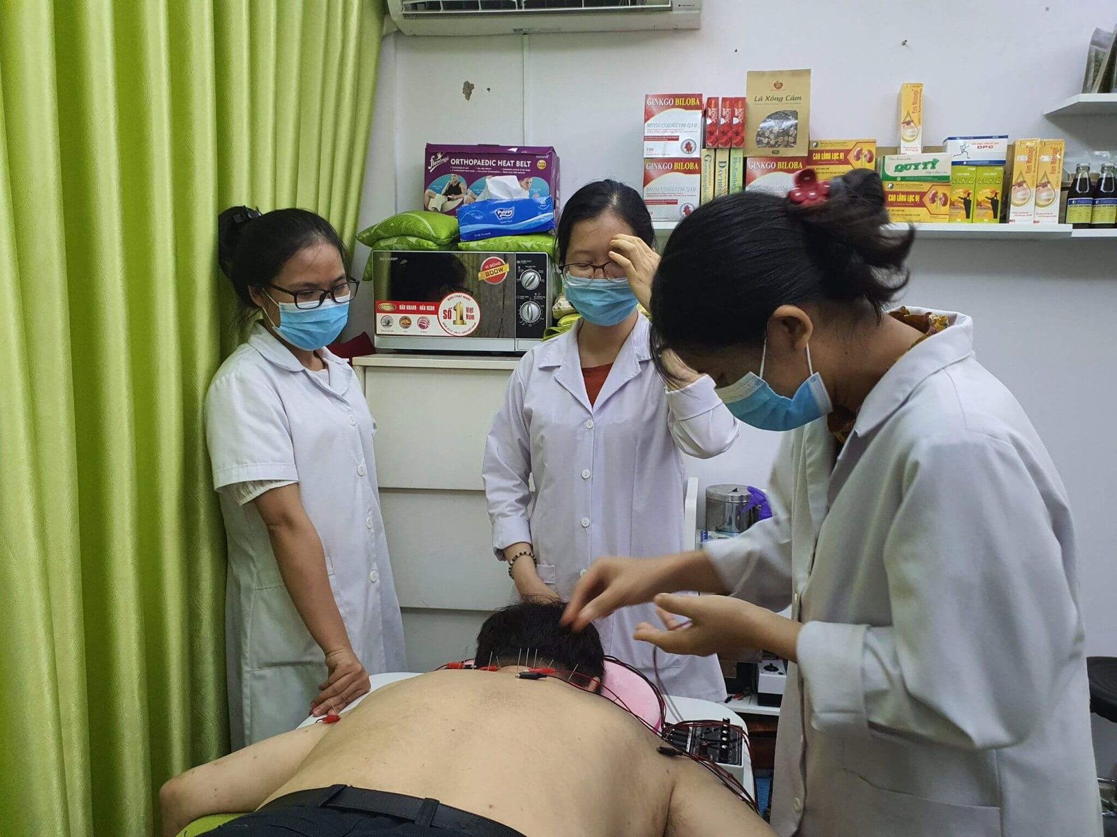Ba người mặc áo blouse trắng đang thực hiện liệu pháp châm cứu cho một người nằm sấp trong phòng có rèm xanh.