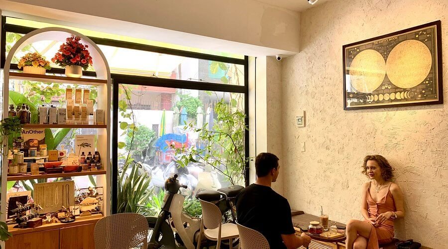 Hai người ngồi uống cà phê trong quán, bên cạnh là kệ sách và cửa sổ lớn nhìn ra phố với cây xanh.
